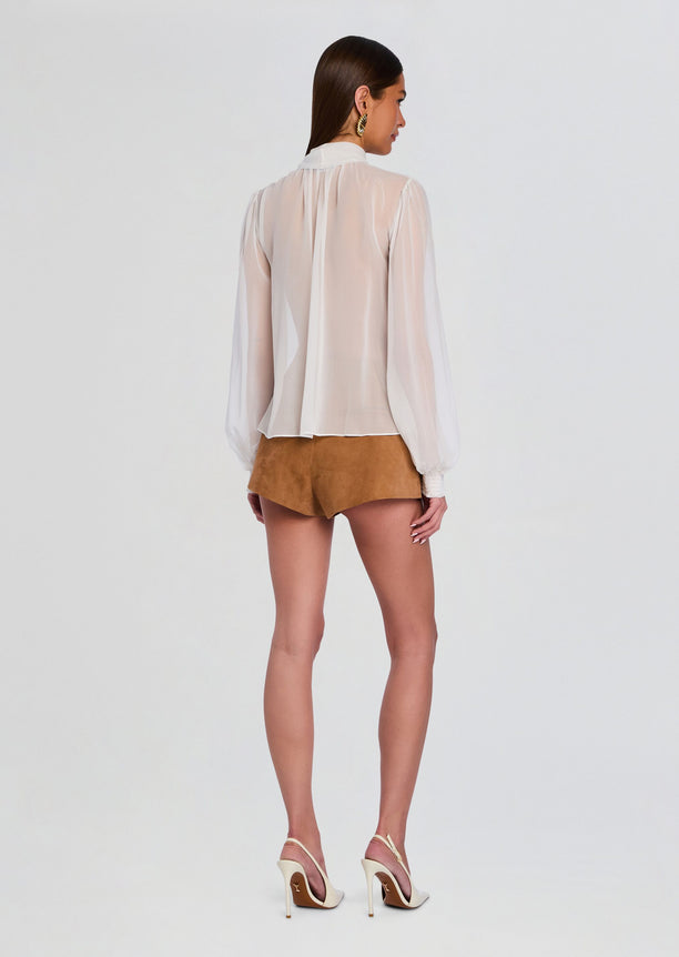 Back view of Hasli Silk Chiffon Top on model, sheer long sleeve blouse with gathered back and smocked cuffs, worn with tan shorts.