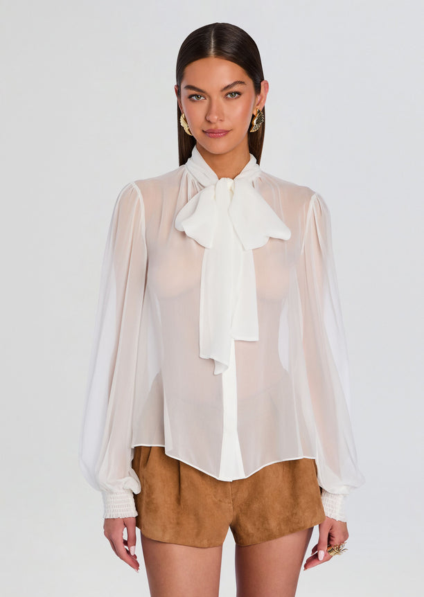 Front view of Hasli Silk Chiffon Top with large bow at the neck, sheer puff sleeves, paired with camel suede shorts.