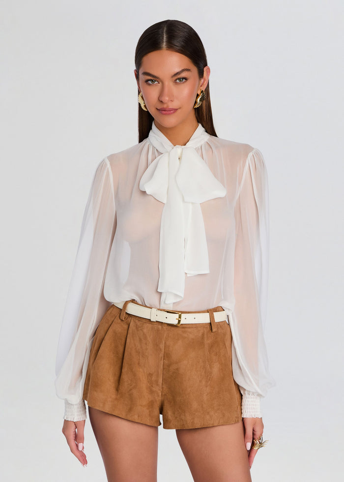 Model wearing white sheer silk chiffon top with a large neck bow, tucked into camel suede shorts. media 1 of 6