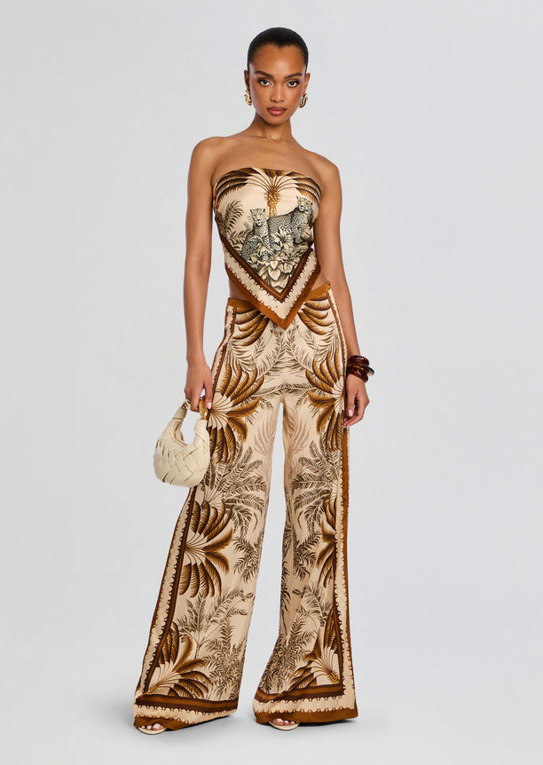 Model wearing the Sunny Pant set with a strapless bandeau top and matching wide-leg pants in beige and brown palm print, with a scarf belt and cream bag.