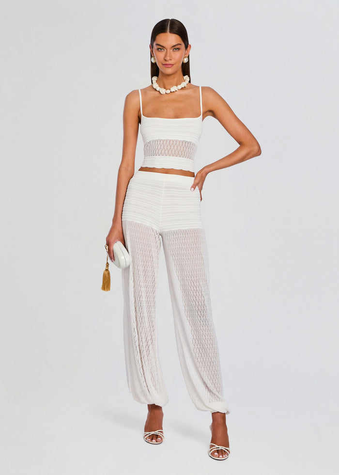 Model wearing a white crochet crop top with a lattice waist detail and matching sheer pants, holding a white clutch. media 2 of 5