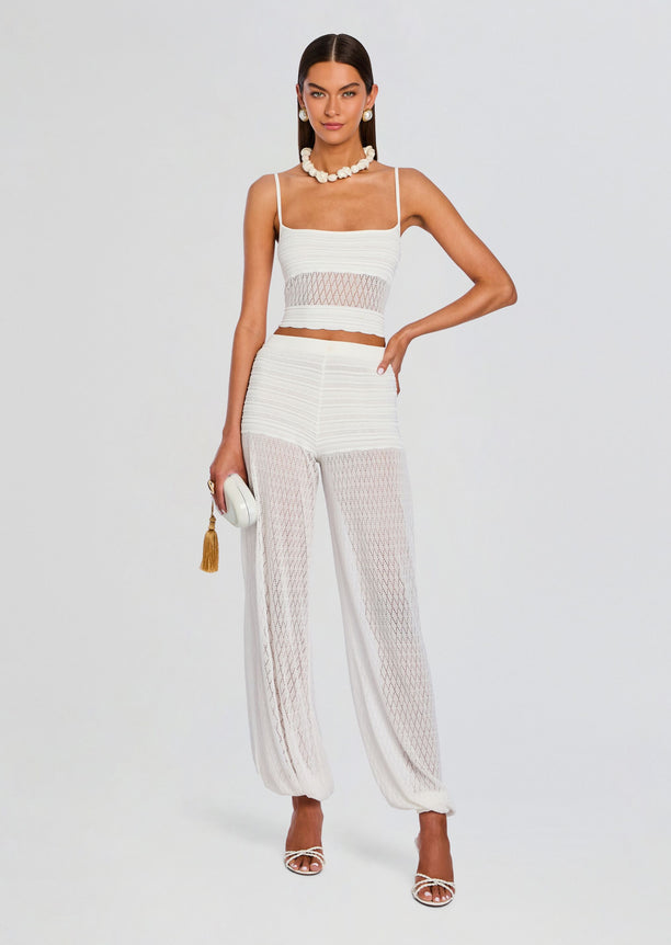 Model wearing a white crochet crop top with a lattice waist detail and matching sheer pants, holding a white clutch.