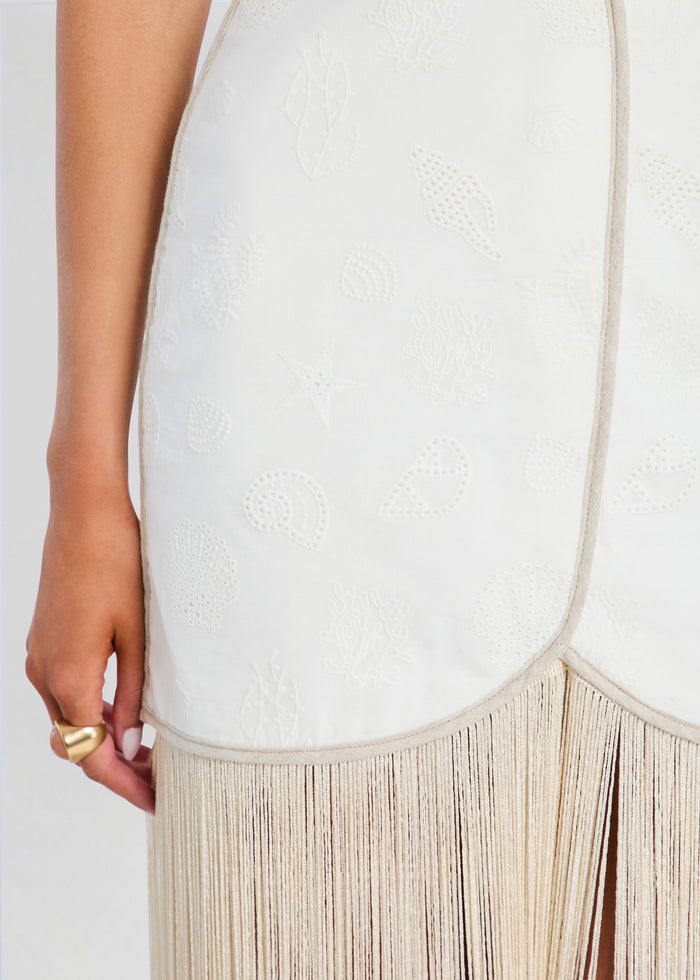 Close-up of a cream-colored embroidered dress with a beige fringe hem, worn by a model whose hand and arm are visible on the left side. media 2 of 5