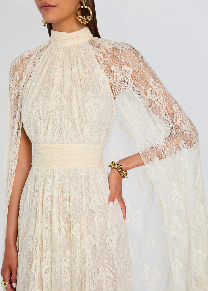 An off-white lace dress with sheer cape sleeves, a high mock neck, and a matching fabric waistband, shown on a model against a light gray background. media 2 of 5