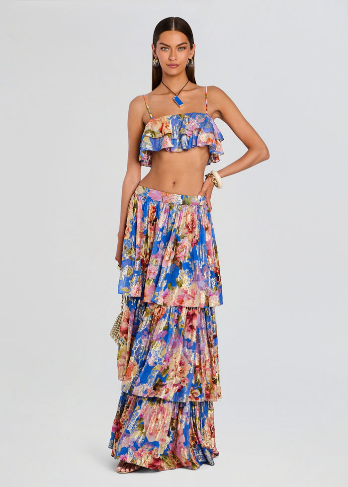 A model wearing a vibrant, multicolored floral print two-piece set consisting of a ruffled crop top and a matching tiered maxi skirt, photographed against a solid light gray background. media 2 of 5