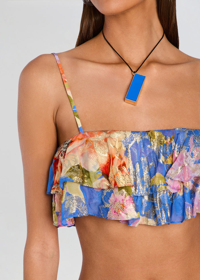 A close-up of a model wearing a ruffled strapless top with a vibrant blue, pink, and gold abstract floral print, paired with a geometric blue pendant necklace. media 2 of 5