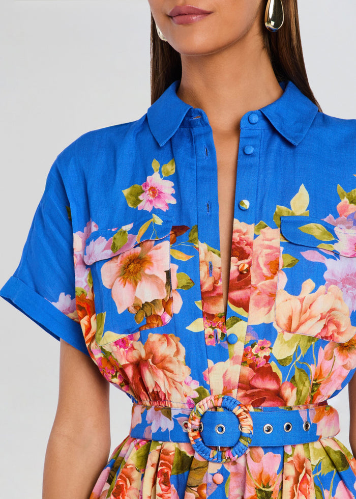 A close-up of a model wearing a bright blue short-sleeve collared shirt dress with a colorful floral print and a matching blue belt with a round woven buckle. media 2 of 5