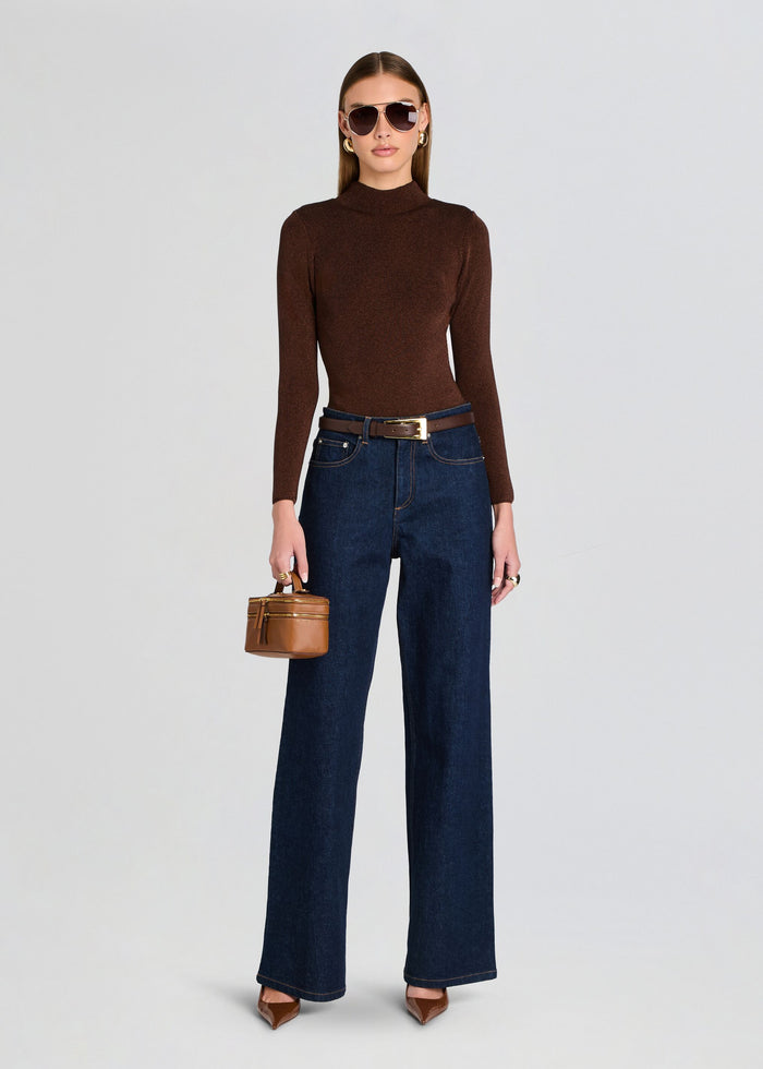 Woman wearing a brown sweater and dark blue jeans, holding a small brown handbag on a light gray background. media 2 of 5