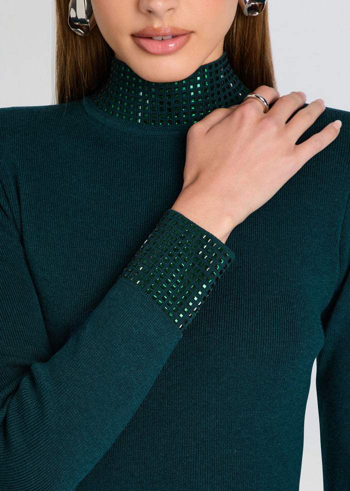 A close-up of a model wearing a dark green ribbed knit mock neck sweater with sparkling green sequined trim at the high collar and cuff. media 2 of 5