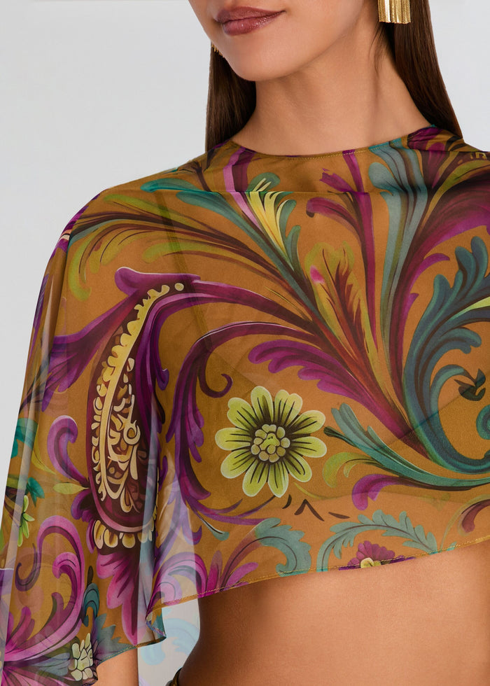 A close-up of a model wearing a sheer, semi-transparent crop top with a vibrant paisley and floral print in purple, teal, gold, and green on a mustard background. media 2 of 5