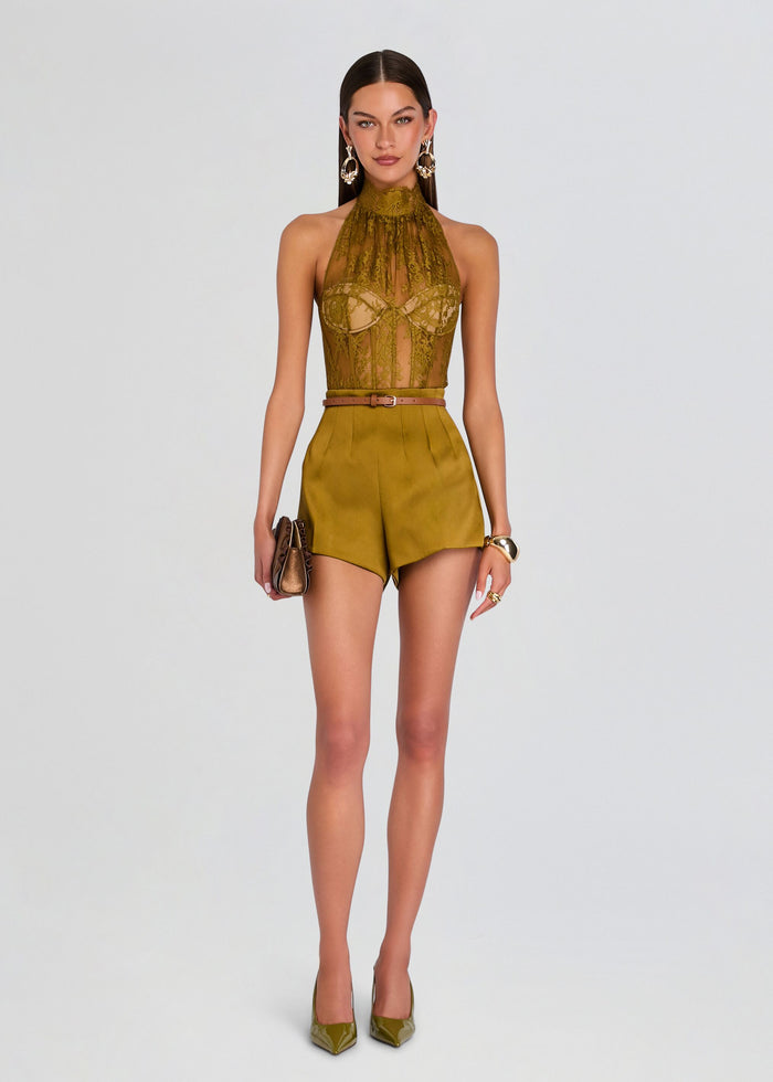 A model stands against a light gray studio background wearing a matching olive green two-piece outfit consisting of a sheer lace halter top and high-waisted shorts, paired with a thin brown belt and olive green pointed-toe heels. media 2 of 5