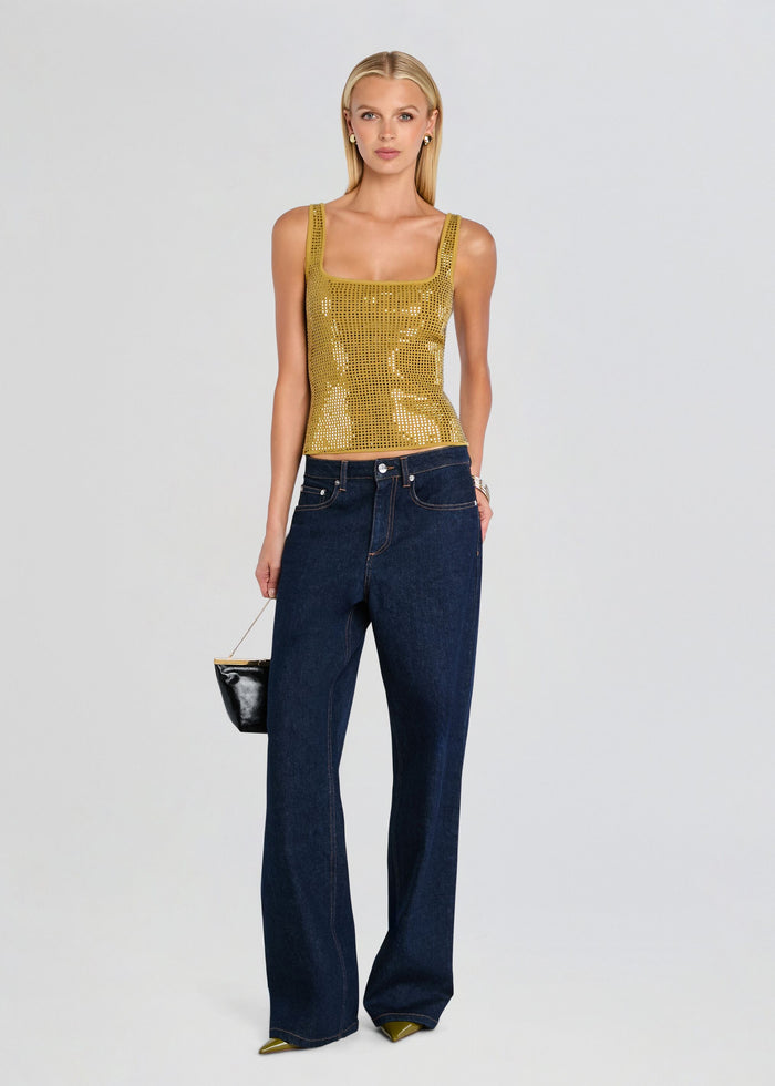 A blonde model stands against a light gray studio background wearing a gold sequined sleeveless top and dark blue wide-leg jeans, holding a small black handbag. media 2 of 5