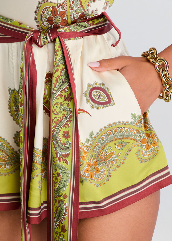 Close-up of a printed skirt or dress with a paisley pattern in olive green, yellow, burgundy, and cream, featuring a matching tie belt and a pocket with a hand inserted. media 2 of 5