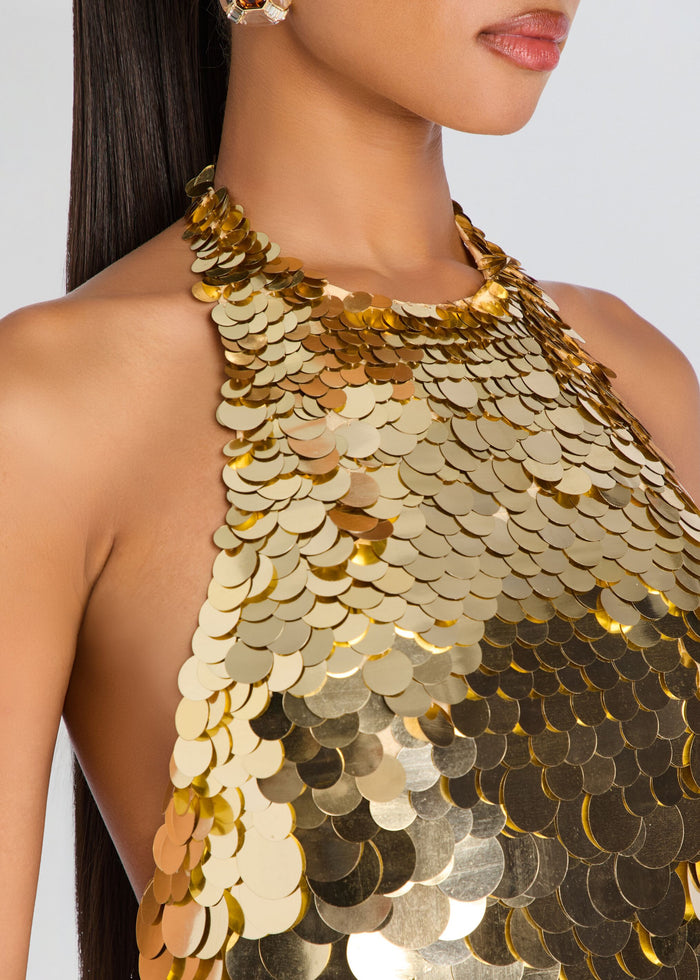 Close-up of a model wearing a high-neck halter top covered in large, overlapping metallic gold sequins against a light gray background. media 2 of 5