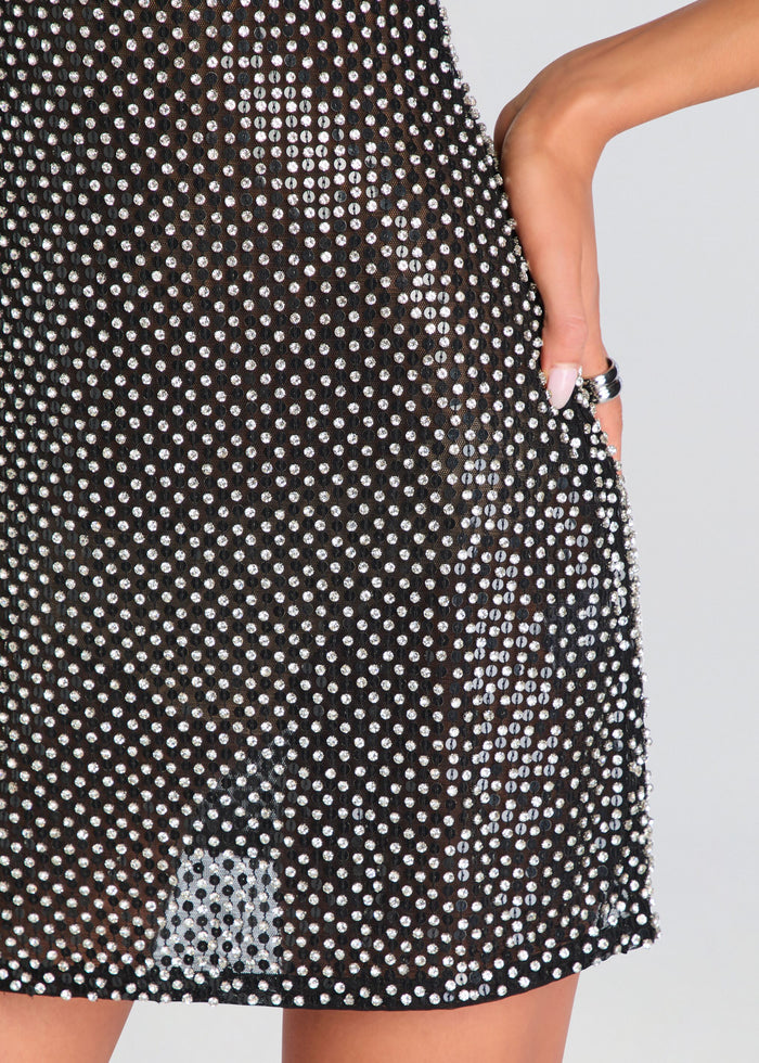Close-up of a black sheer mesh dress covered in small silver rhinestones, worn by a model with one hand in the pocket, against a light gray background. media 2 of 5