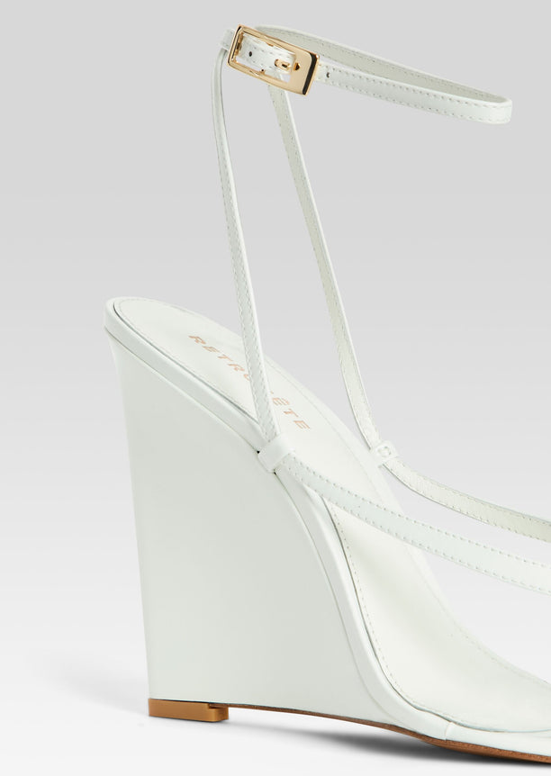 Close-up of Namby Wedge white sandal showing ankle strap with gold-tone buckle and wedge heel