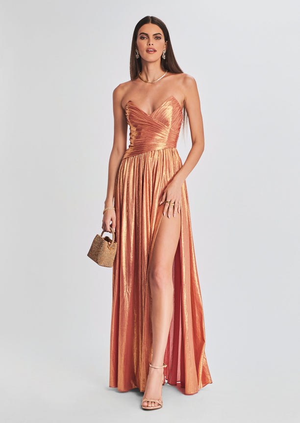 Waldorf Dress in metallic rose gold featuring a ruched bodice and flowing skirt with a high slit, model wearing statement earrings.